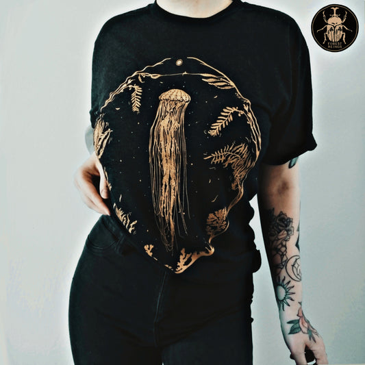 Cute witchy goth girl with long black hair and tattoos on her arms and legs, wearing an aesthetic goth graphic t-shirt. The print on the t-shirt is a big golden jellyfish and an oval frame, full of seaweed and other marine fauna. 