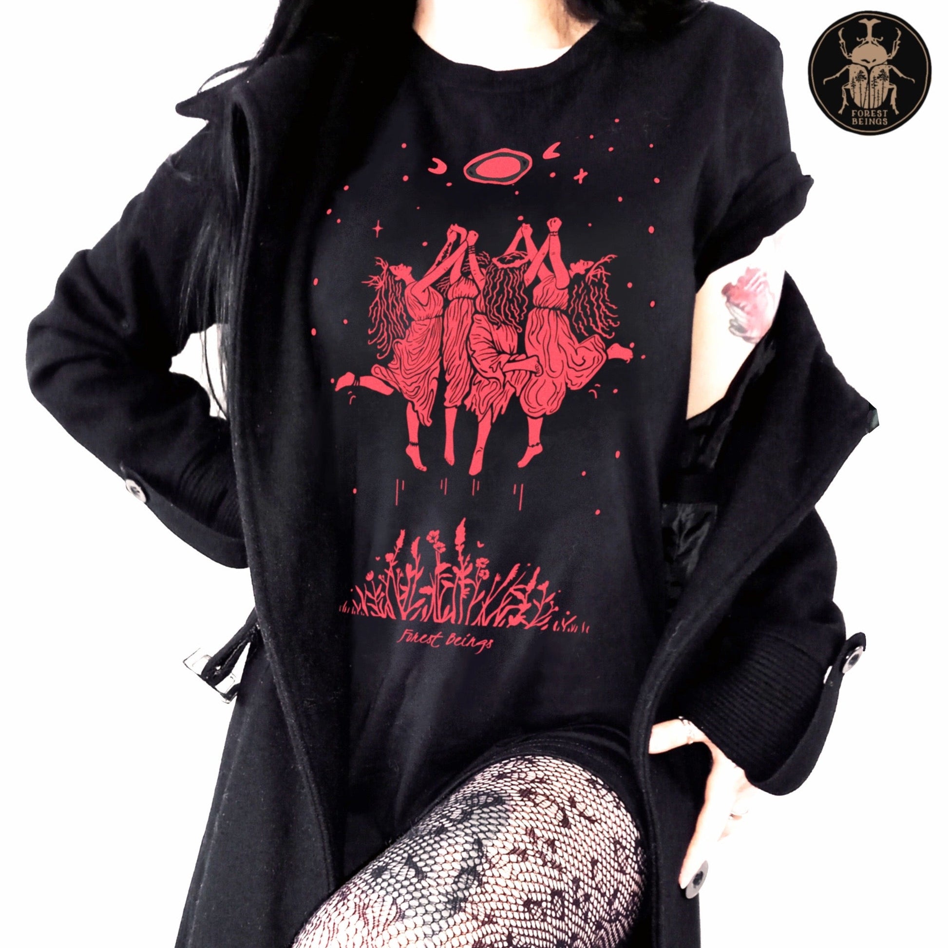 Witchy graphic t-shirt where witches are dancing in the air, in a sabbath ritual like act. Cute witchy goth girl with long black hair and tattoos on her arms and legs, wearing an aesthetic goth graphic t-shirt. 
