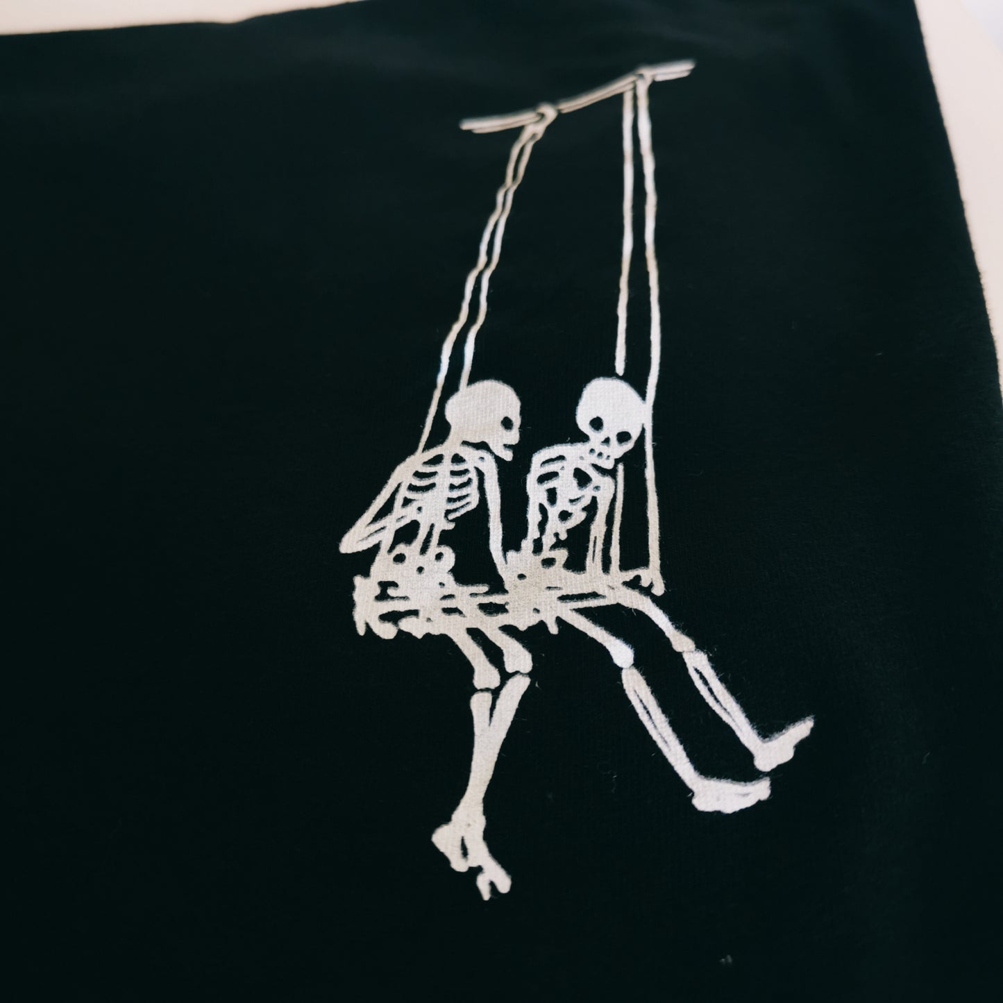 Two white skeletons on a swing, screen printed on a pocket of a gothic t-shirt. 