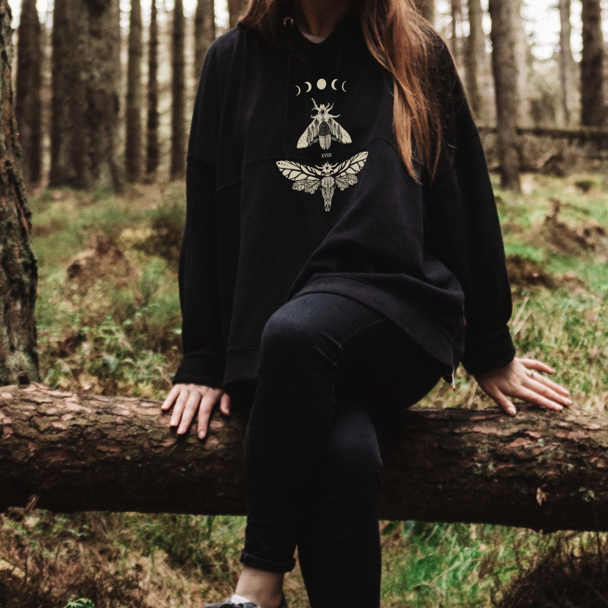 A gothic girl, in the forest, is wearing a black moths witchcore oversized hoodie. 