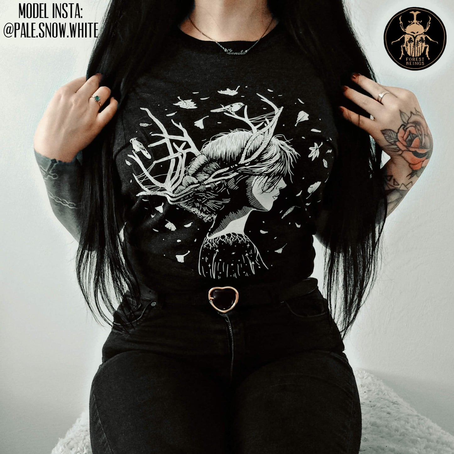 Cute witchy goth girl with long black hair and tattoos on her arms and legs, wearing an aesthetic goth graphic t-shirt. The design of the t-shirt is a forest witch, surrounded by falling autumn leaves. 
