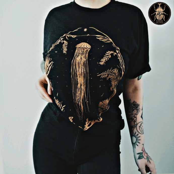 Cute witchy goth girl with long black hair and tattoos on her arms and legs, wearing an aesthetic goth graphic t-shirt. The print on the t-shirt is a big golden jellyfish and an oval frame, full of seaweed and other marine fauna. 