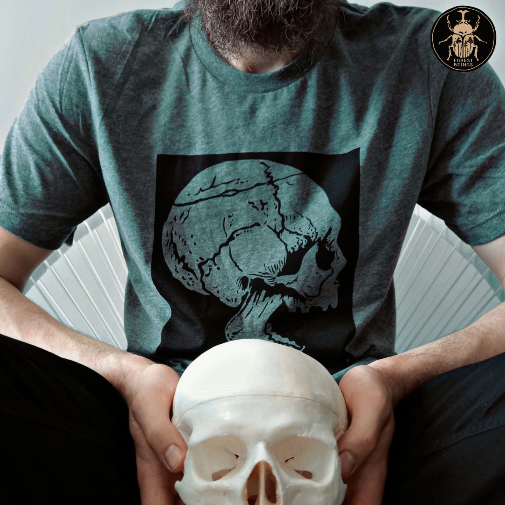 A gothic guy with a beard, holding a skull and wearing a t-shirt with a skull print on it.