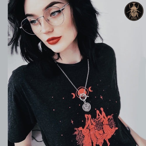 Cute witchy goth girl with long black hair and tattoos on her arms and legs, wearing an aesthetic goth graphic t-shirt. The print on the t-shirt is red witches sabbath, where witches are levitating and dancing in the air. 